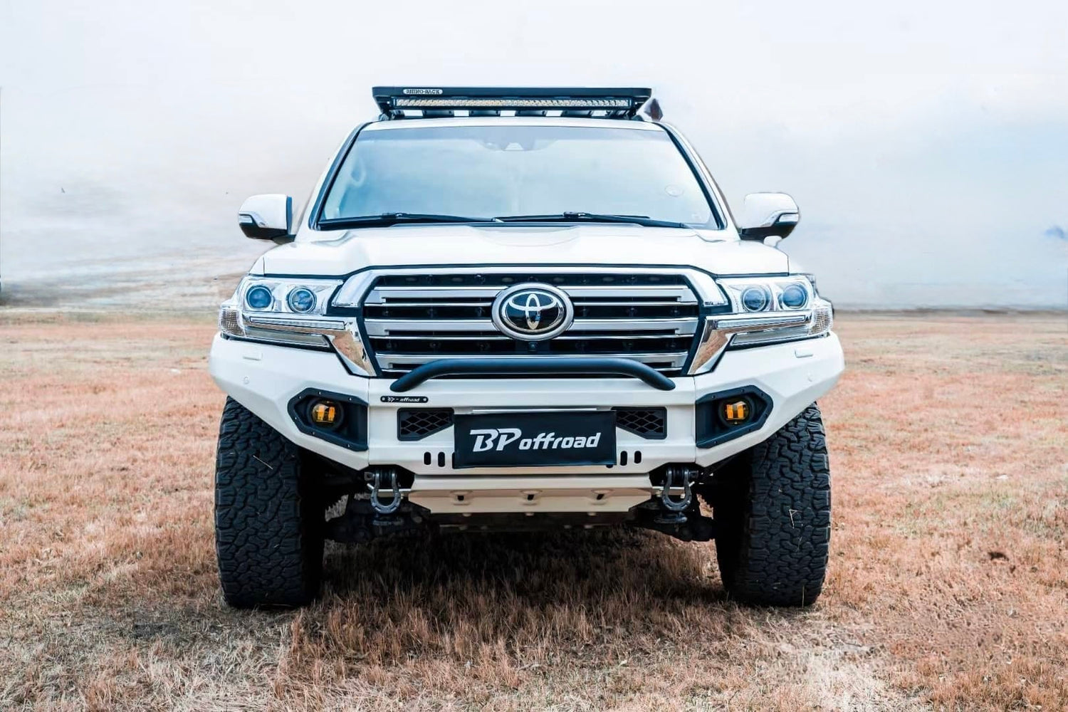 white suv toyota land cruiser lc200 with heavy duty white powder coated front bumper fitted 2 yellow fog lights and roof rack led light bar on dry grass pavement