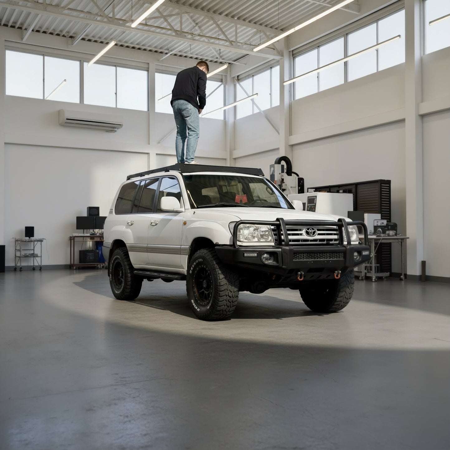 White Land Cruiser 100 series in a large indoor space with a person standing on top of the modular slim roof rack- sherpa style